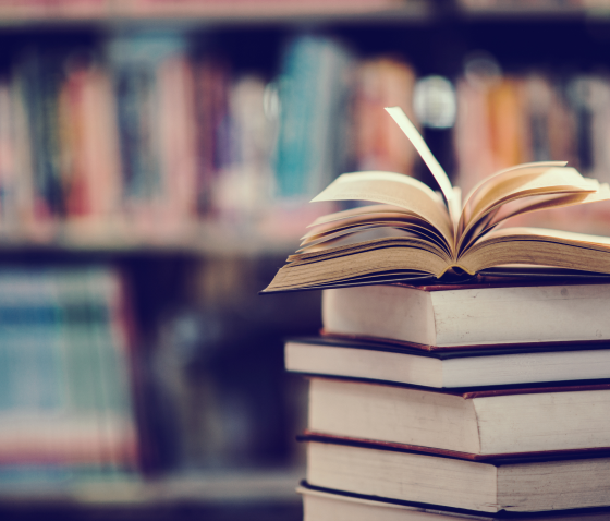 Pilha de livros com um livro aberto no topo, em uma biblioteca, simbolizando conhecimento, aprendizado e pesquisa
