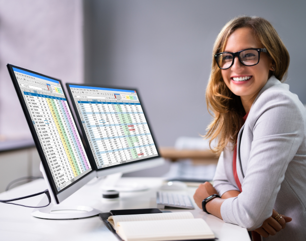 Analista financeira sorridente trabalhando com planilhas em dois monitores, representando análise de dados e gestão empresarial